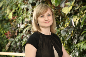 A blond woman wearing a black, short-sleeved dress and white pearl necklace standing in front of a wall of greenery.
