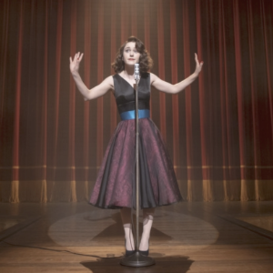 A screenshot from a TV show: a young woman in 50's style dress and hair standing in front of a microphone on a stage.