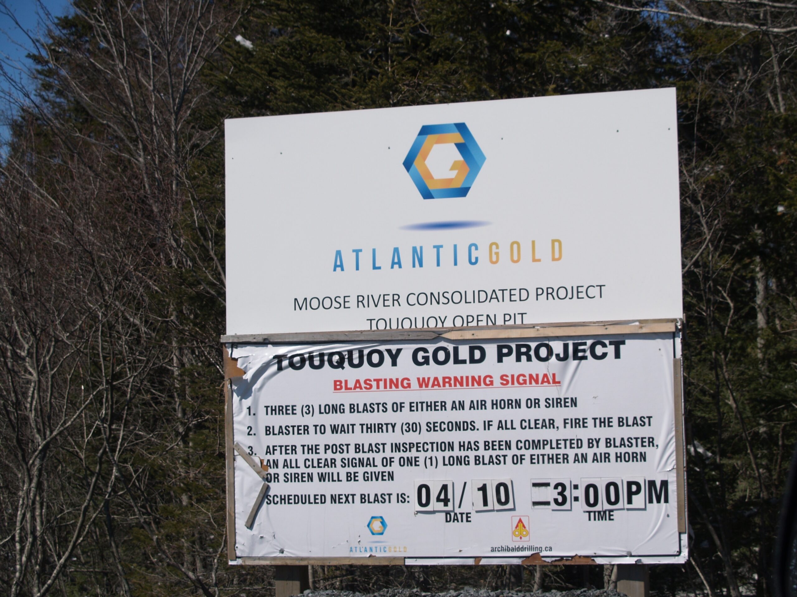 这张2018年在驼鹿河金矿现场的广告牌显示，该矿山是驼鹿河联合项目的一部分，还有大西洋黄金公司的标志和名称，在广告牌下面是Touquoy黄金项目的每日爆破时间表，警告每个人当天爆破将在什么时间发生。照片:琼巴克斯特