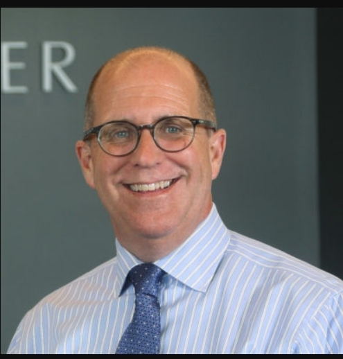 A balding man with glasses wearing a pale blue shirt with blue tie