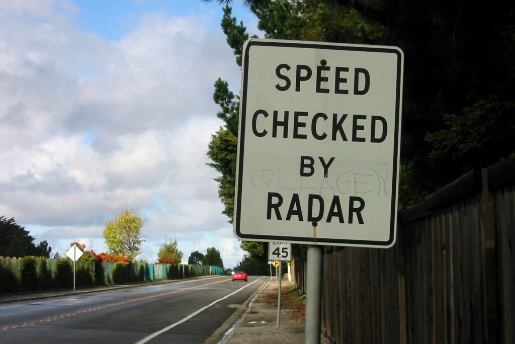 路边有一个写着“雷达检测速度”的标志。在那个标志后面是另一个标志，表示限速45。背景中有一辆红色的汽车。夏天或秋天是阴天。