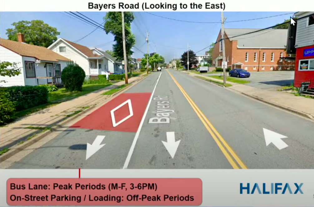 A Google Streetview image shows a road annotated with arrows. On the left side, there are two lanes pointing in the direction of the viewer. One of those lanes, the furthest left, has a red section with a white diamond, denoting a bus lane. There is one lane on the right, leading away from the viewer. On either side of the street are buildings, houses on the left and a church and storefront on the right. It's a sunny day without a cloud in the sky, and the foliage is green.