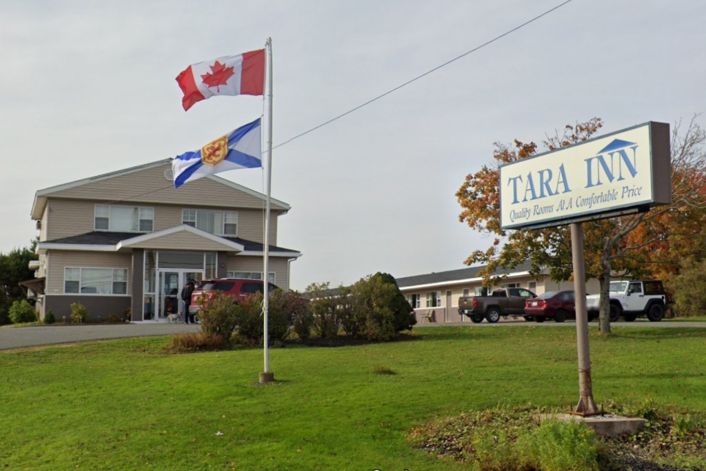 A motel is seen on an overcast day. In the front lawn, there's a flag pole with the Canadian and Nova Scotian flags flying. In front of the flag pole is a sign reading, TARA INN Quality Rooms At A Comfortable Price.