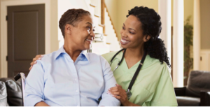 一名身穿浅绿色磨砂风格上衣、脖子上挂着听诊器的微笑女子，正把手放在一名身穿浅蓝色衬衫的老妇人肩上，对她微笑。