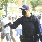 一名身份不明的警察戴着黑色的帽子，戴着墨镜和面具，咄咄逼人地拿着一罐胡椒喷雾