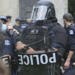 A very intimidating Halifax police officer, dressed in full black gear with a padded vest, black helmet, visor, police shield and baton.