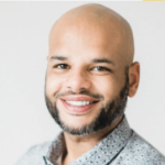 A smiling young Black man with a shaved head and trim beard