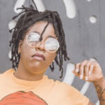 A young Black woman with round sunglasses and an orange T shirt