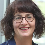A white woman with wavy dark chin length hair and dark rimmed glasses