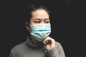 An Asian woman in a beige turtleneck sweater wearing a blue disposable mask against a black background