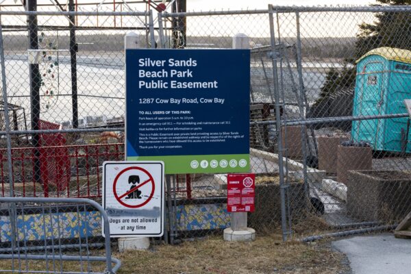 可以看到杂乱的标识和围栏。最大的标志是蓝绿相间的，白色的字母写着公园的名字和地址，牛湾道1287号。下面是一个划掉狗的牌子，上面写着:“现在任何时候都可以带狗进来。”中间有一个移动厕所，后面是海滩。