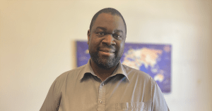 身穿灰领衬衫的黑人对着镜头微笑，背景墙上的世界地图模糊了