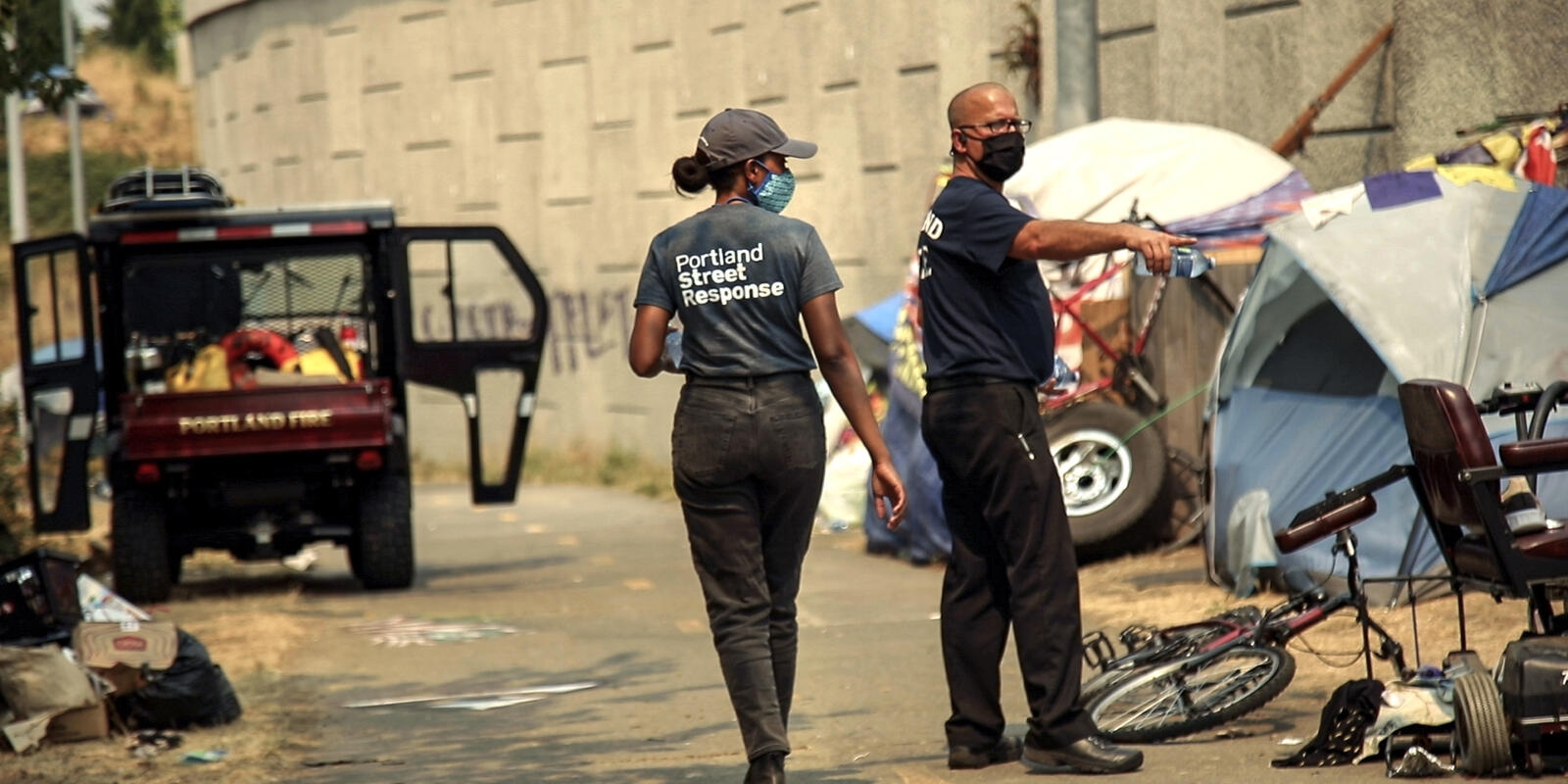 一个黑人男人和一个黑人女人，都戴着面具，站在一个帐篷营地的小巷里。他们穿着写着波特兰街头反应的t恤