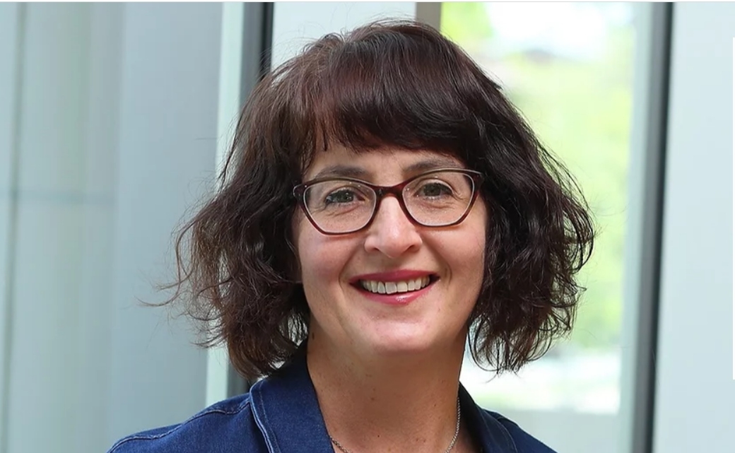A woman with a wavy bob haircut, dark rim glasses, wearing a denim jacket.