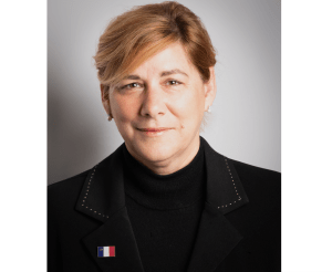 一个微笑的女人，金发短发，穿着黑色衬衫和黑色运动夹克，在她的右翻领上有一个鲜艳的阿卡迪亚国旗别针。