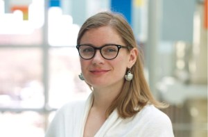 一位戴着眼镜、身穿白衬衫、面带微笑的金发女郎站在一扇明亮的窗户前，阳光透过窗户射进来。