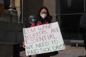 A woman wearing a mask holds a sign that says low workers are essential but we need paid sick days.