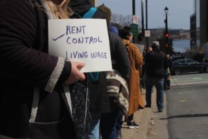 A line of protestors with one holding a sign that says rent control, living wage