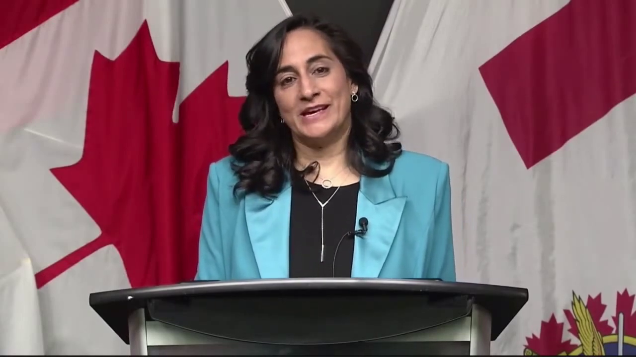 深色长发、淡蓝色西装外套的女士在讲台上讲话，背景是加拿大国旗