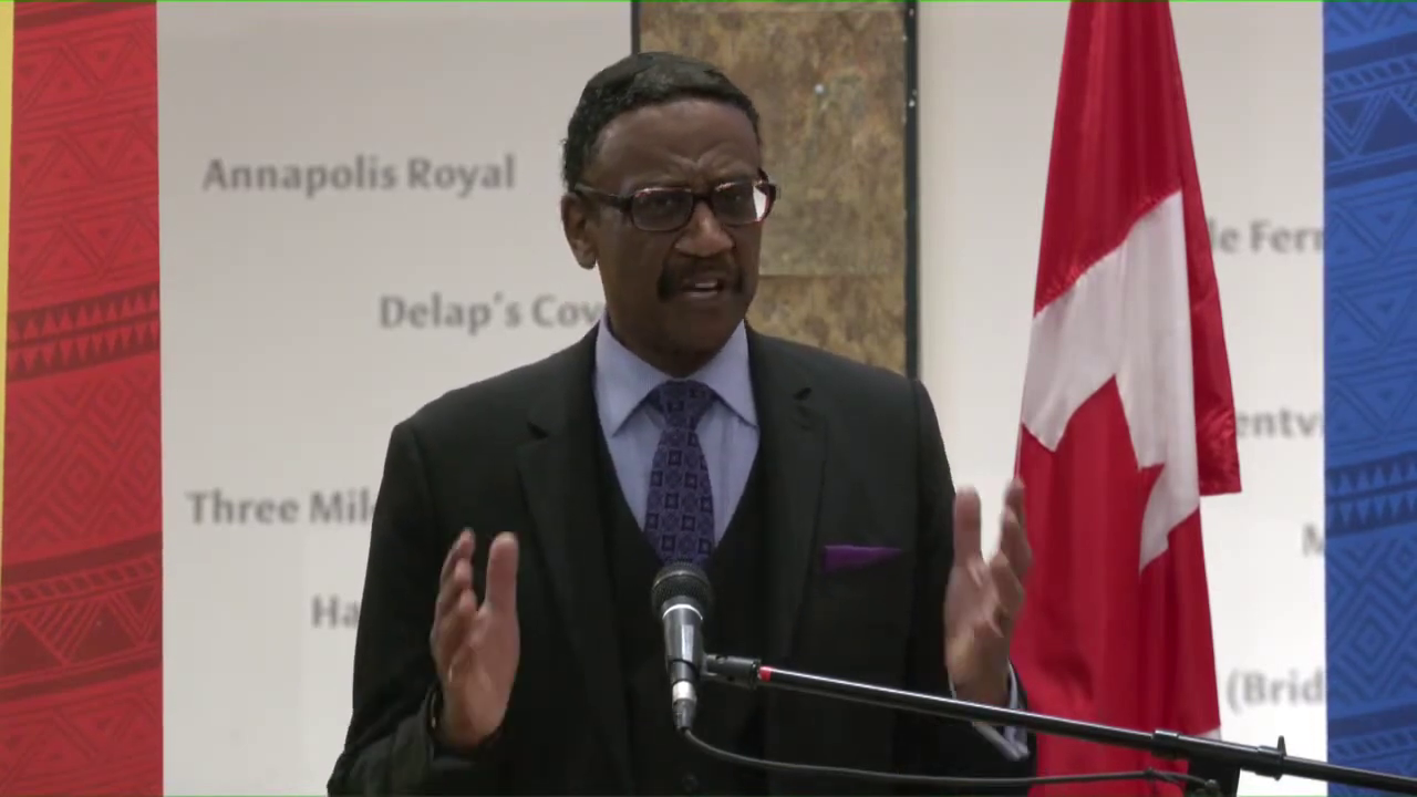 一名戴眼镜、身穿深色西装的黑人男子在讲台上发表演讲，背景是加拿大国旗。