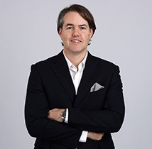 身穿黑色夹克和白色衬衫的中年白人男子双手交叉站着，看着镜头。