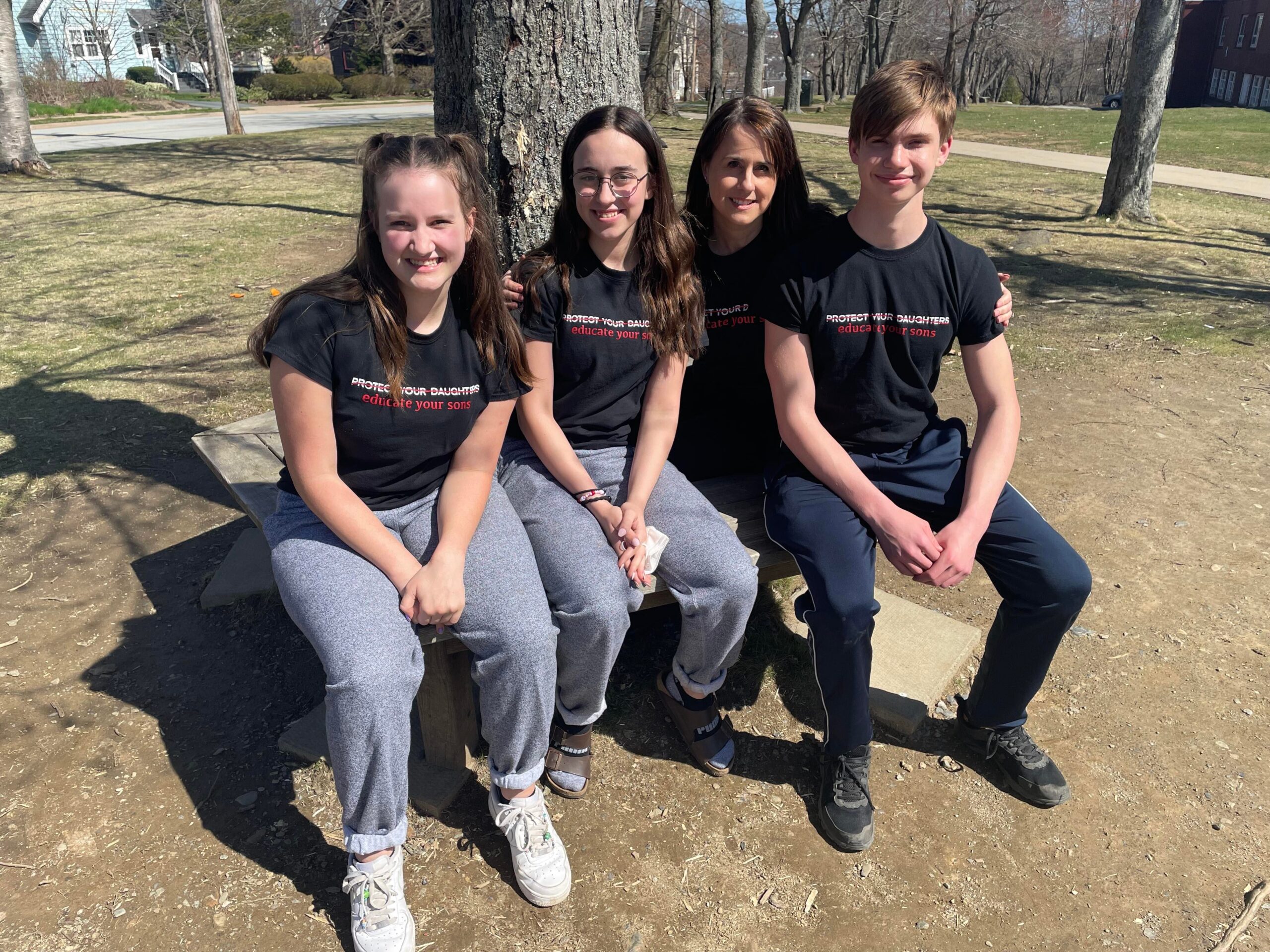四个人坐在一棵树的长凳上。每个人都穿着带有徽标的黑色T恤，上面写着保护您的女儿，这些女儿与一条线路交叉。下面说教您儿子的短语。