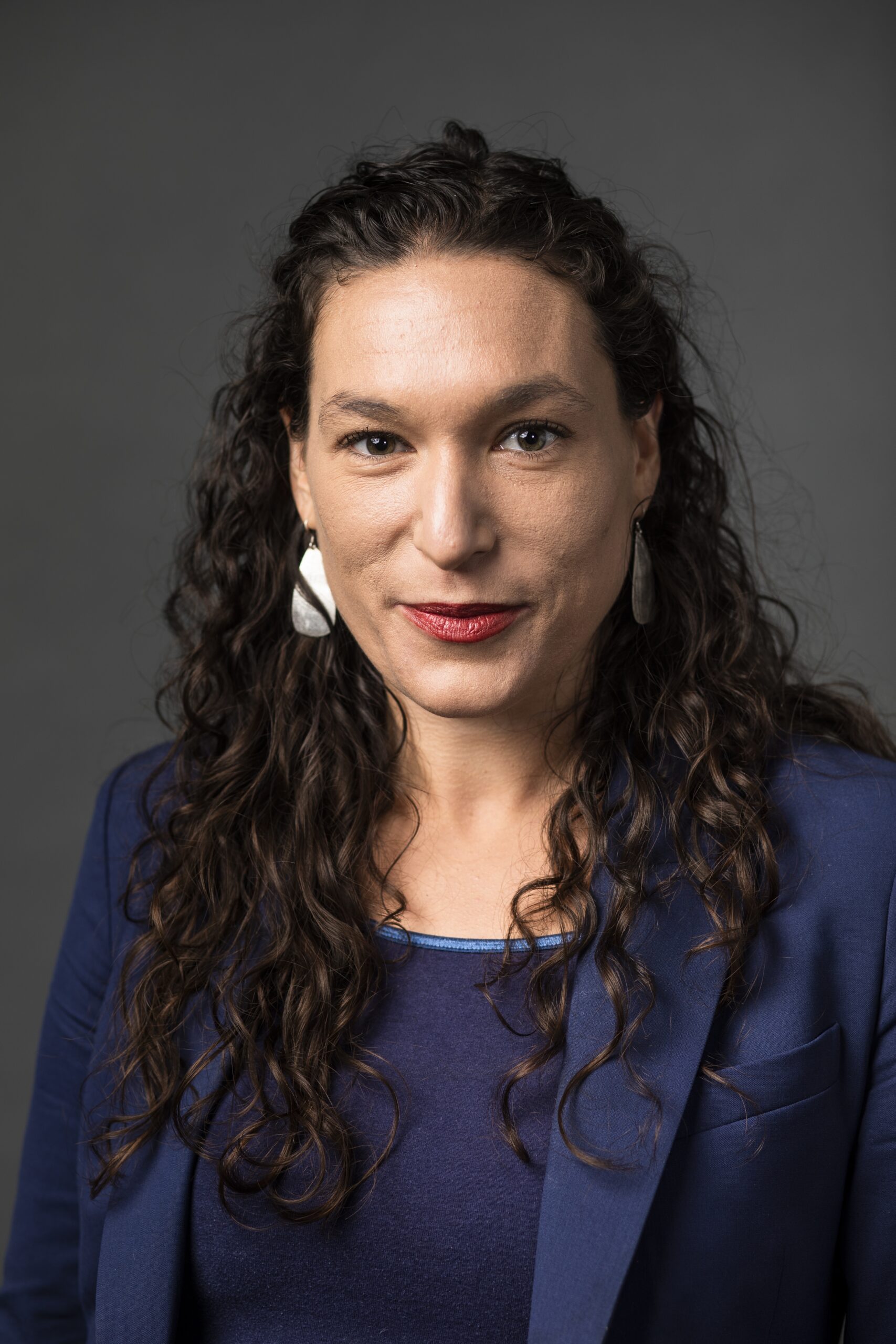 一个白人女人的头像，长卷发，戴着白色大耳环。她穿着蓝色上衣和蓝色运动夹克，直视镜头。