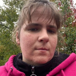 一名戴着耳环、黑色毛衣外搭粉色毛衣的女子看着镜头。在她的身后是树上的绿色和红色的叶子。