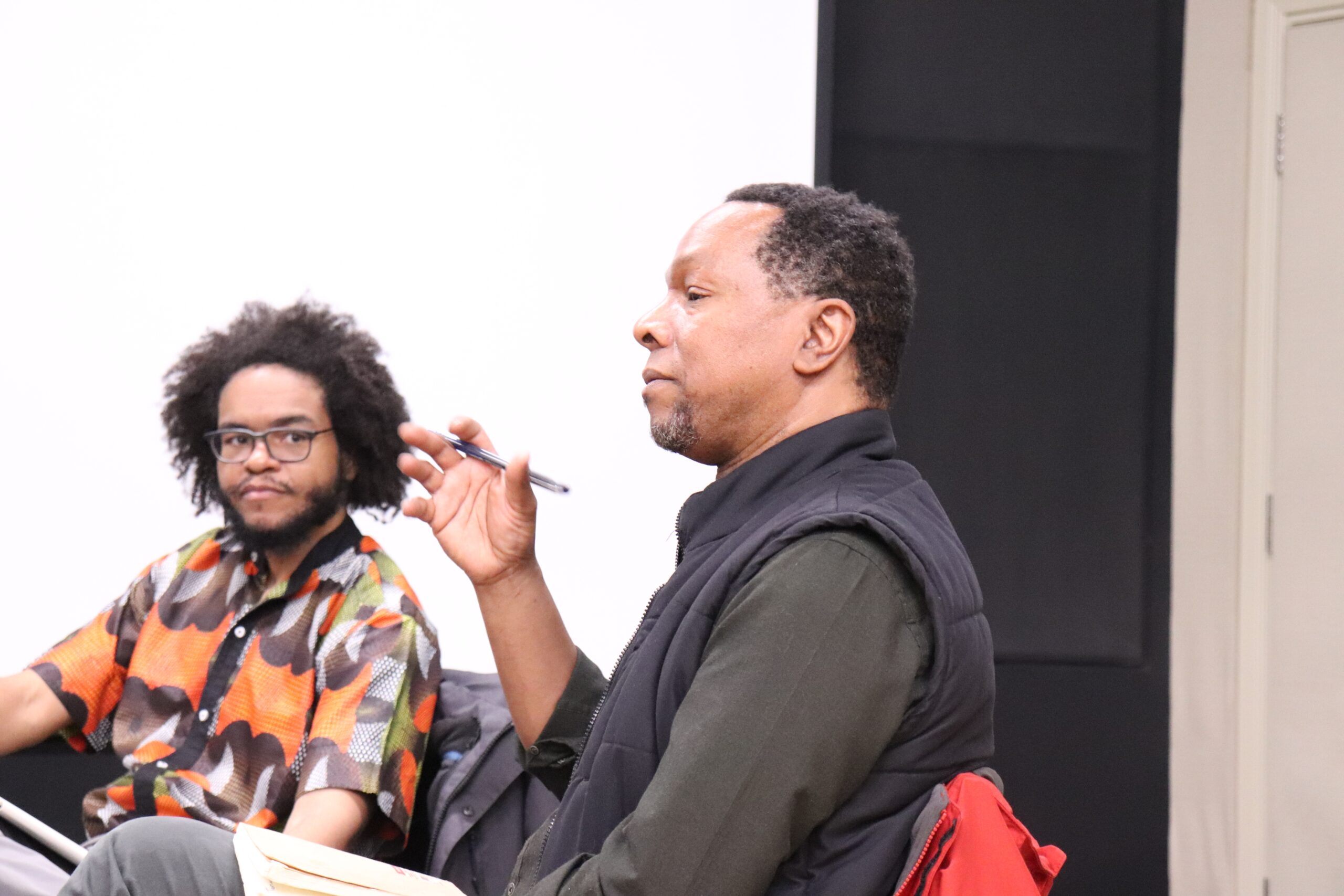 Black man with afro and glasses and multi coloured shirt and black men holding a pen in long sleeve dark green shirt and black vest.