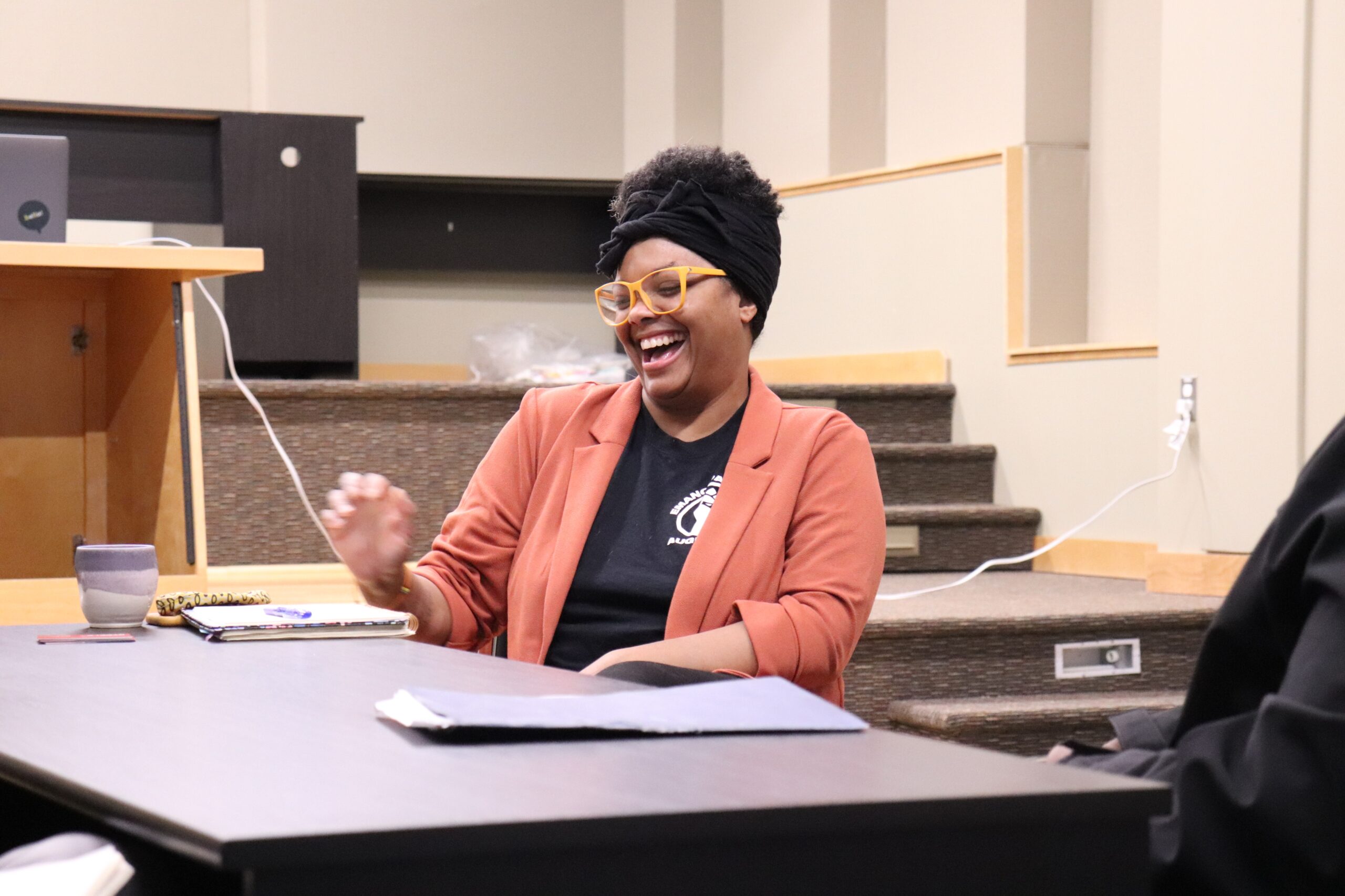 Black lady with yellow glasses and orange blazer smiles and laughs