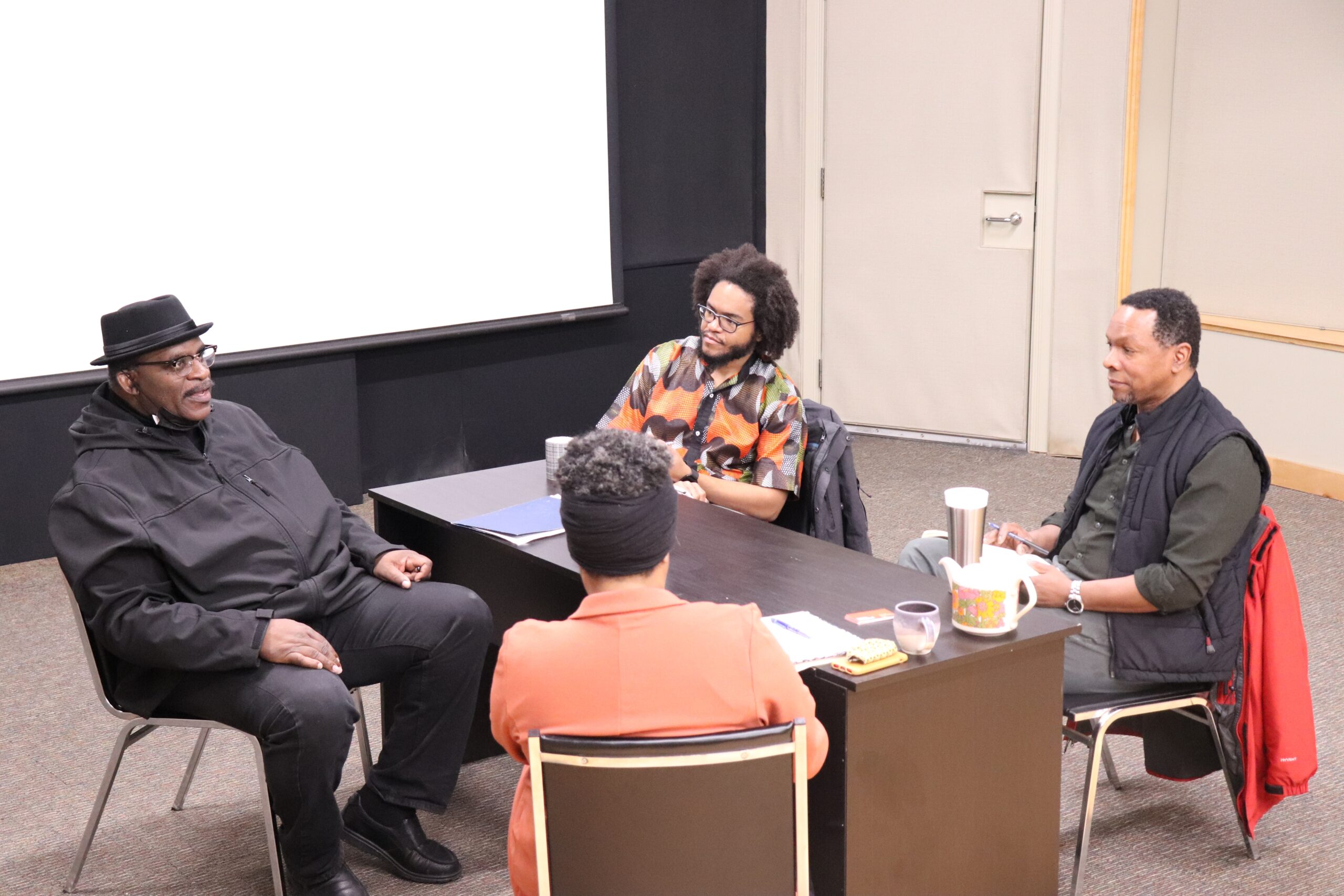 Three Black men sit around a table with a black woman who's back is to the camera