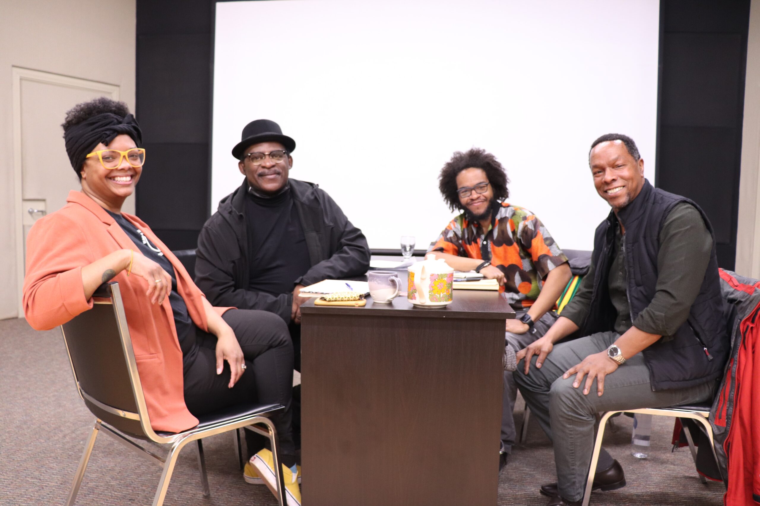 黑人妇女和三个Lack男子对着镜头微笑，围坐在一张桌子旁，背景是白色的屏幕。