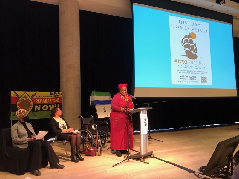 身穿红衣服、戴着红头巾、戴着眼镜的黑人女士在讲台上讲话