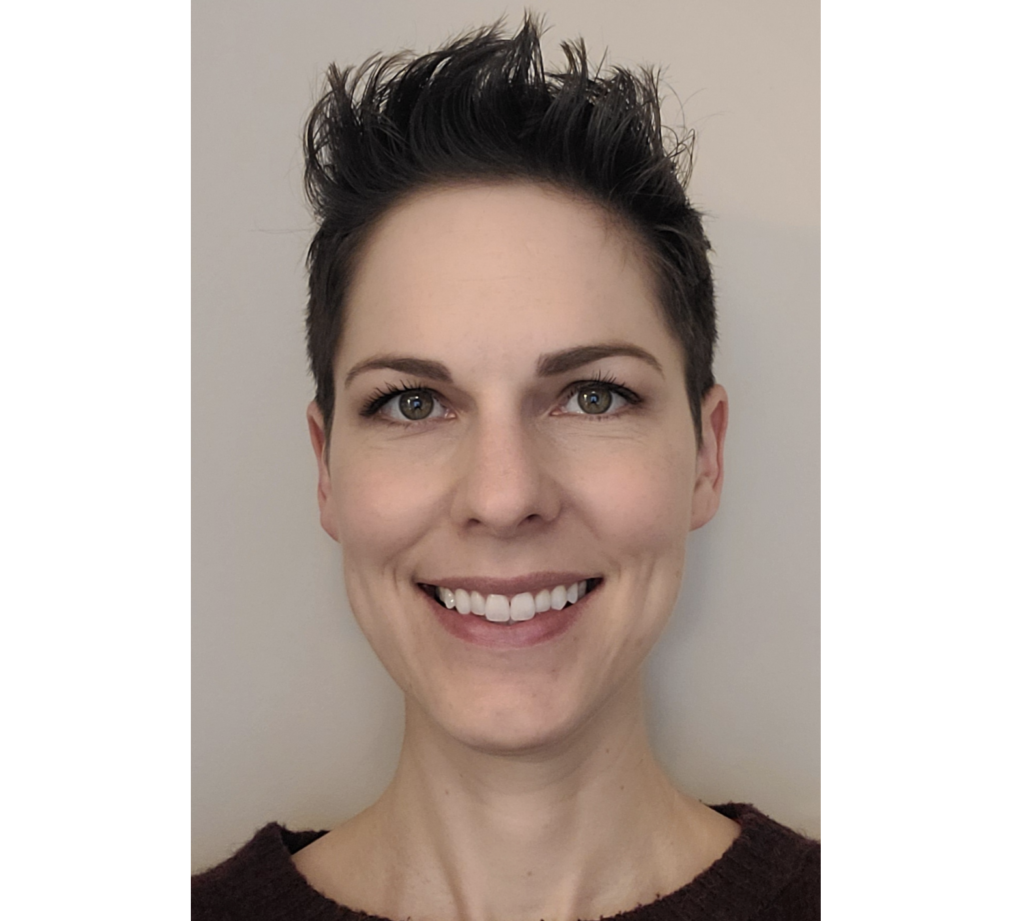 一个微笑的女人,短的他ar wearing a dark top and standing against a beige wall smiles at the camera.