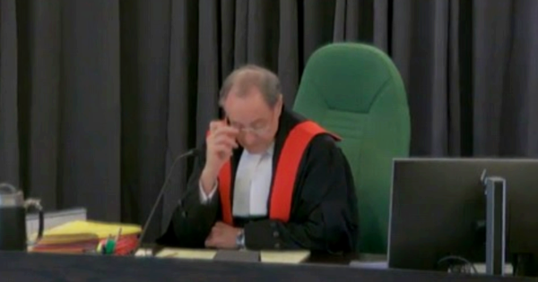 Judge Warren Zimmer, an older white man with grey hair, wearing black robes with a red border, reads from documents at his desk.