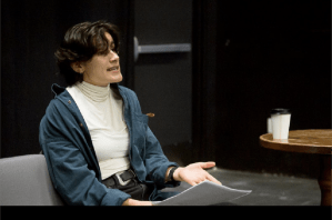 一个留着深色波浪发型的女人，穿着蓝色开衫和白色高领毛衣，坐在桌旁读稿子。