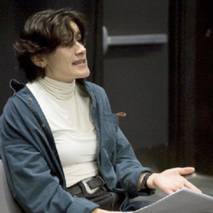 A white woman with short dark wavy hair wearing a blue cardigan discusses a script