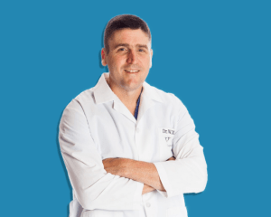 一个穿着白色白大褂和蓝色手术服的白人抱着胳膊站着。