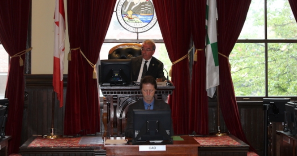 Peter Kelly at a Charlottetown city council meeting, sitting at a large desk with a small sign reading 