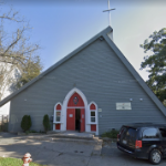 a grey A frame building with a red door and cross on top
