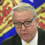 在新闻发布会上，一位白发苍发、戴着黑框眼镜的老年白人男子站在几面新斯科舍省国旗前