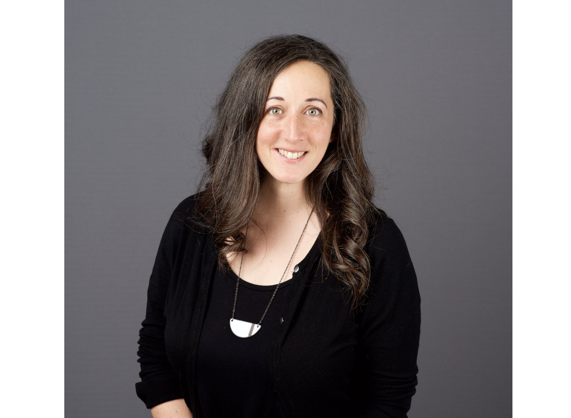 A smiling woman wearing a long sleeved black dress top stands against a dark grey background leaning slightly forward.