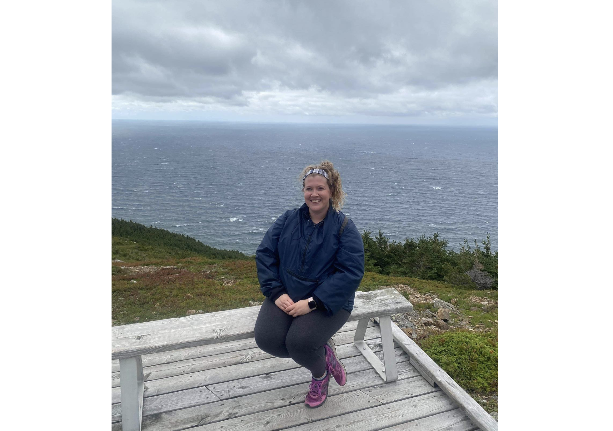 穿深蓝毛线衣和裤子的一个少妇坐一个白色长凳在一个木平台上，横渡的腿，微笑用她的在她的膝部的手。灰色的天空和海洋在她身后。