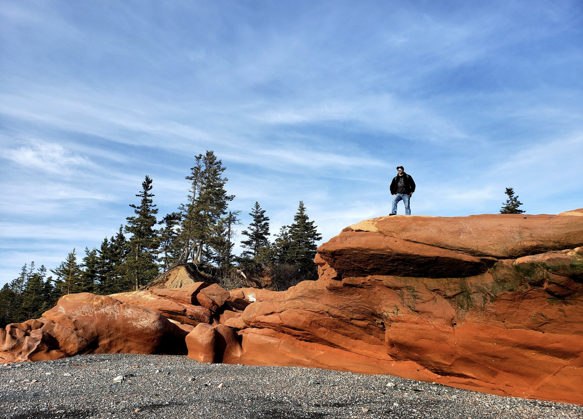 A man is dwarfed standing atop a giant reddish orange rock on the coast with a blue sky and trees behind, a sandy beach below.