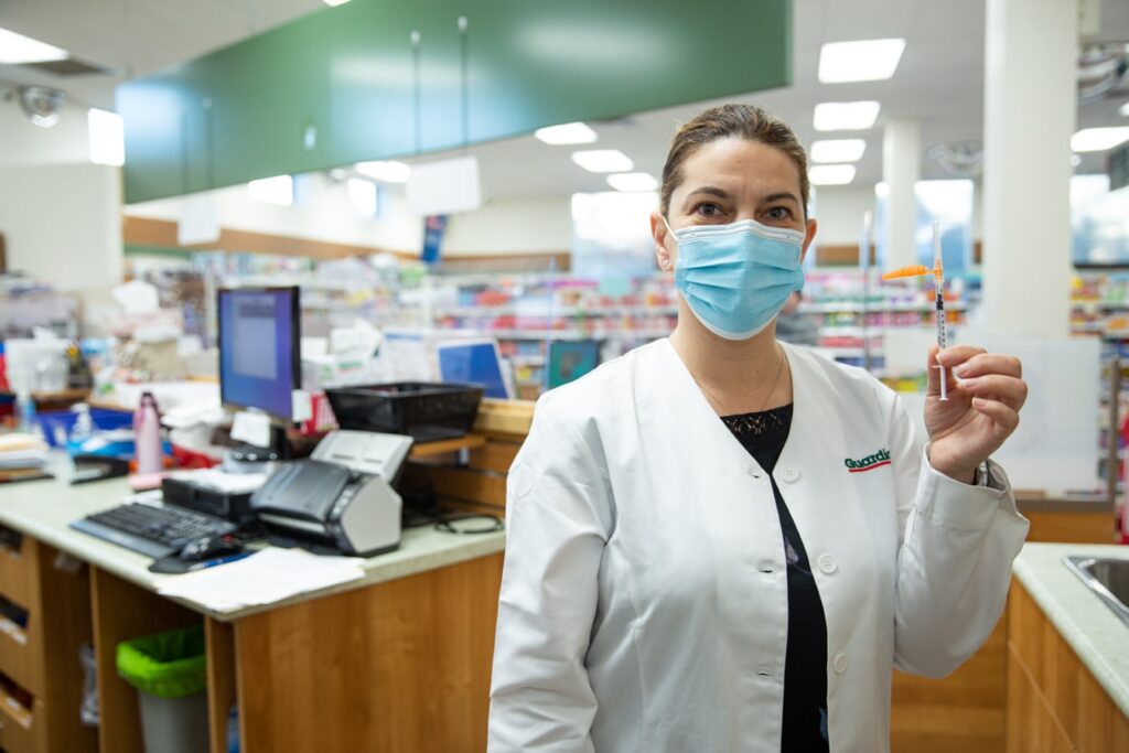 A masked pharmacist stands at her work station holding a needle.