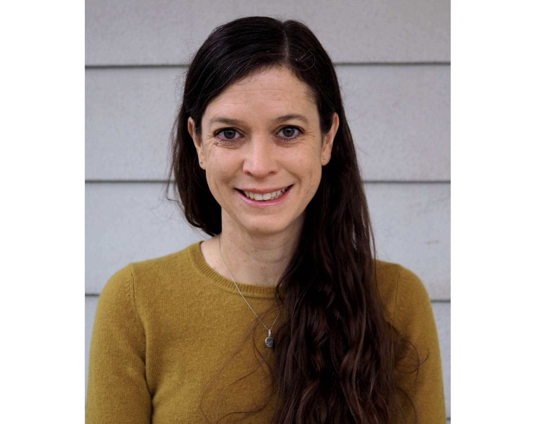 在浅灰色的背景前，一名身穿淡黄色衬衫、黑发长发的微笑女子对着镜头微笑。