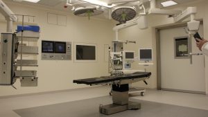 在明亮的灯光和操作设备下，空的手术室里有一张空床。在照片的右边可以看到医生的手。