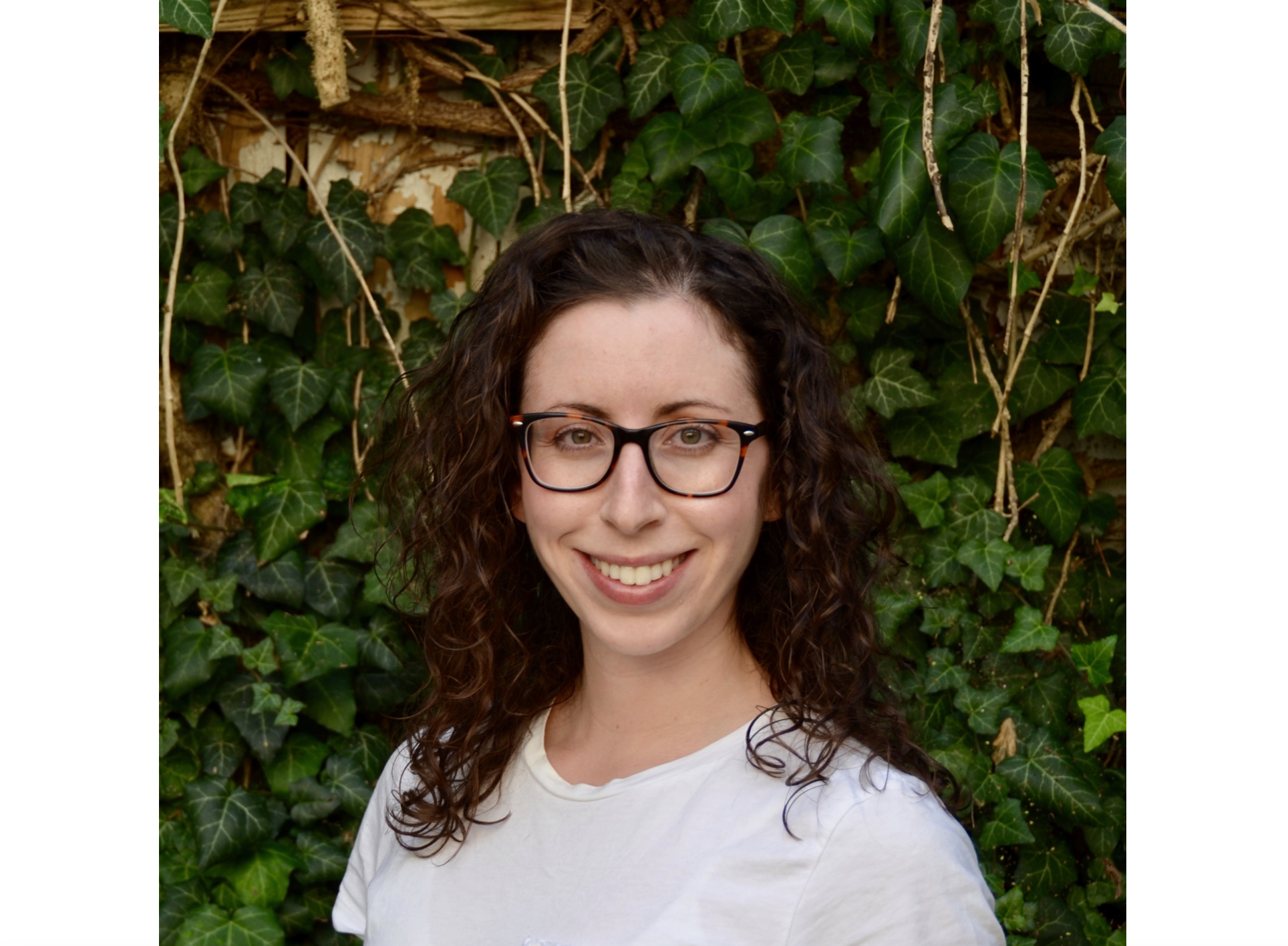 A smiling young woman with curly dark hair and dark glasses and wearing a white top stands against a wall of greenery.