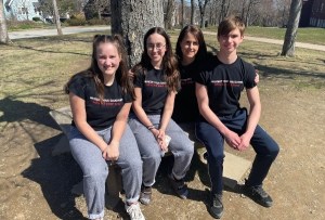 三个白人青少年学生和他们的老师坐在长凳上，穿着相配的黑色t恤