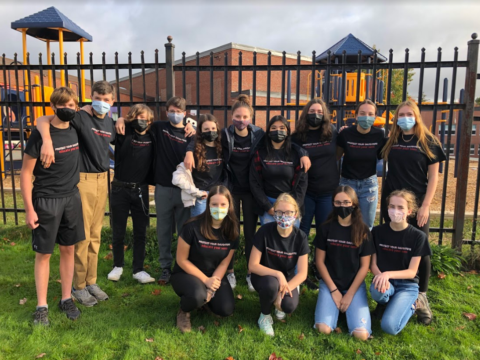 一群穿着黑色T恤的青少年说“保护您的女儿”，其中有一条线，然后在下面写下“教育您的儿子”一词。学生们戴着口罩，站在黑色铁栅栏前的草地上。